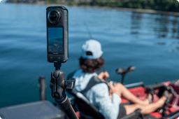 A 360-degree action camera mounted on a black adjustable arm in the foreground, focused on a seated person in a red kayak wearing a light blue jacket and backward cap. The camera captures the serene blue water of a lake with distant tree-lined shores.