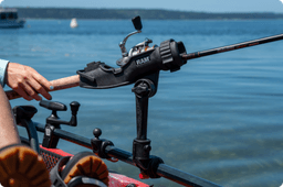Close-up of a RAM brand fishing rod holder mounted to the side of a red kayak, securely holding a fishing rod. A person’s hand adjusts the rod while sitting barefoot in the kayak, with calm lake water and distant boats in the background.