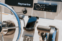 Smartphone secured in RAM® X-Grip® holder mounted on boat helm near Raymarine electronics, providing convenient access to mobile navigation and communication while operating marine vessel.