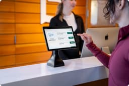Visitor interacting with a self-service check-in kiosk at a reception desk. A tablet mounted on a desktop stand displays a digital visitor management interface with options for guest registration and check-in. A receptionist stands behind the counter while the visitor selects an option on the touchscreen, illustrating front desk operations, visitor management, self-service registration, and customer engagement in corporate, healthcare, hospitality, and commercial environments.