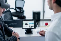 Customer and sales representative reviewing product information on a tablet-based kiosk at a service counter. The tablet is mounted on a desktop stand and displays a product catalog with images, specifications, and purchasing details. Both individuals are focused on the screen during the consultation, illustrating retail, customer service, point-of-sale, and interactive product discovery applications in a commercial environment.