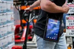 Warehouse worker wearing a rugged tablet in a protective case suspended from a shoulder strap for hands-free mobility. The tablet rests at the worker’s side while they move through a warehouse environment, with palletized inventory, equipment, and safety signage visible in the background. The wearable setup enables quick access to applications, barcode scanning, inventory management, order fulfillment, and warehouse workflows while keeping both hands available for material handling tasks.